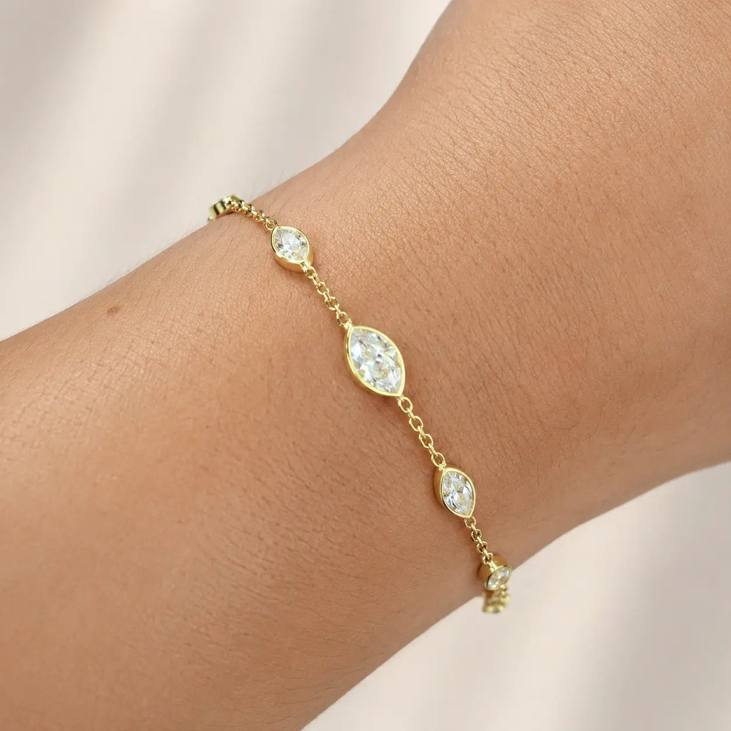 A delicate gold chain bracelet with three marquise-cut clear crystals, shown worn on a person's wrist against a soft beige background.